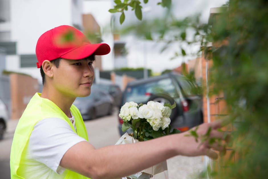 A young male flower delivery courier wearing a red cap and a high-visibility yellow vest gently placing a bouquet of white hydrangeas wrapped in white paper against a hedge outdoors. The arrangement i