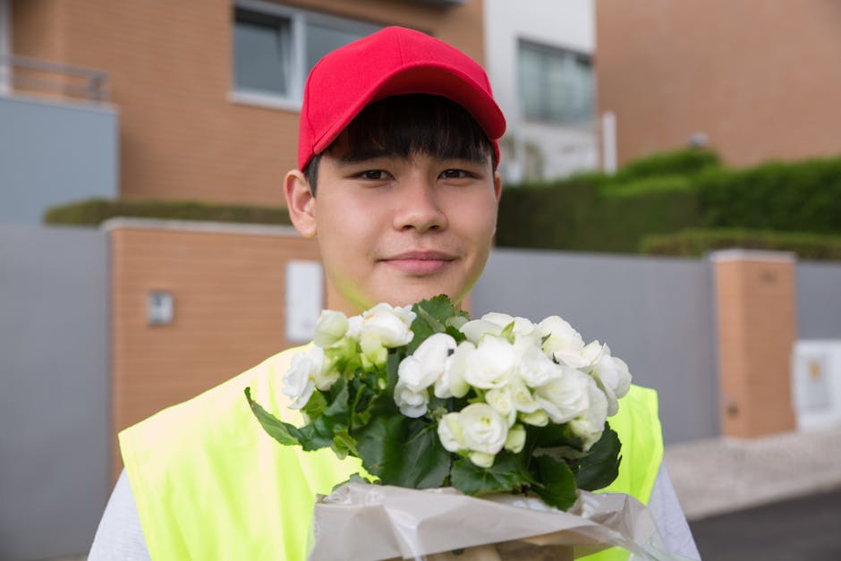 A young male flower delivery courier wearing a red cap and high-visibility yellow vest is standing outdoors in front of modern residential buildings, holding a bouquet of fresh white flowers wrapped i