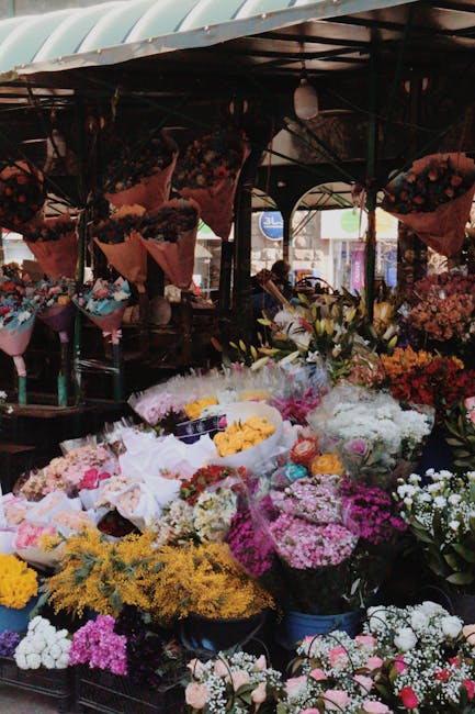 A vibrant floral display at an outdoor market stall, featuring a variety of fresh flowers including white lilies, pink roses, yellow chrysanthemums, purple asters, and multicoloured carnations, arrang