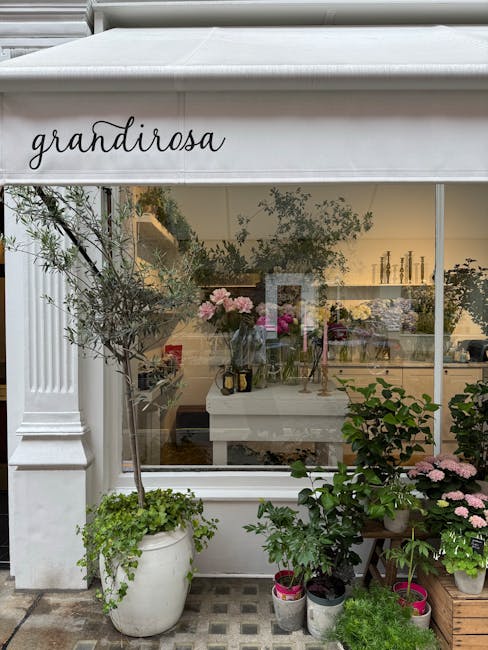 The image shows a florist shop named 'grandirosa' with a large glass window displaying the interior. Outside, there are several potted plants and flowers arranged on the pavement, including a tall oli
