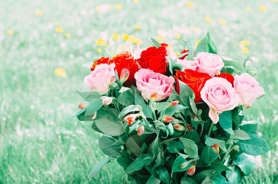 A vibrant bouquet of fresh pink roses, red carnations, and white lilies with orange tips, surrounded by lush green foliage and wrapped in green paper, is held in a person's hand in an outdoor setting