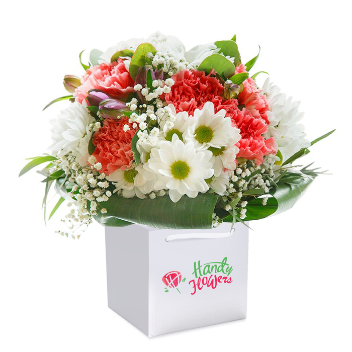 Presented upright in a clean, square white bag, this vibrant bouquet is a testament to floristry skill developed through years serving St Paul's Cray residents. The arrangement bursts with coral and peach carnations, their frilled petals unfurling in luxurious layers, offset by crisp white chrysanthemum daisies whose lime-green centers add a fresh, lively spark. White gypsophila is woven gently throughout, softening the transitions between blooms, while tight purple-hued buds offer moments of anticipation-subtle hints almost hidden, inviting closer inspection like discovering a flowering shrub along a leafy Orpington path. The entire composition is wrapped in a collar of luxuriant, glossy foliage whose depth and sheen serve as the perfect backdrop for the colourful blooms. Light touches every petal evenly, emphasizing the bouquet's rounded fullness and radiant health. Ideal for delivery across St Paul's Cray-from footpaths beside the River Cray to the quiet corners of Crofton Road-this bouquet is perfect for birthdays, anniversaries, or spontaneous tokens of kindness. The white ribbon handles complete this neat yet inviting package, promising a flawless gift that celebrates both tradition and the flourishing of community life.